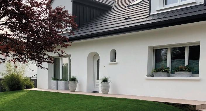 Maison rénovée avec une façade blanche moderne, un toit en ardoise et un jardin bien entretenu avec des plantes en pot.