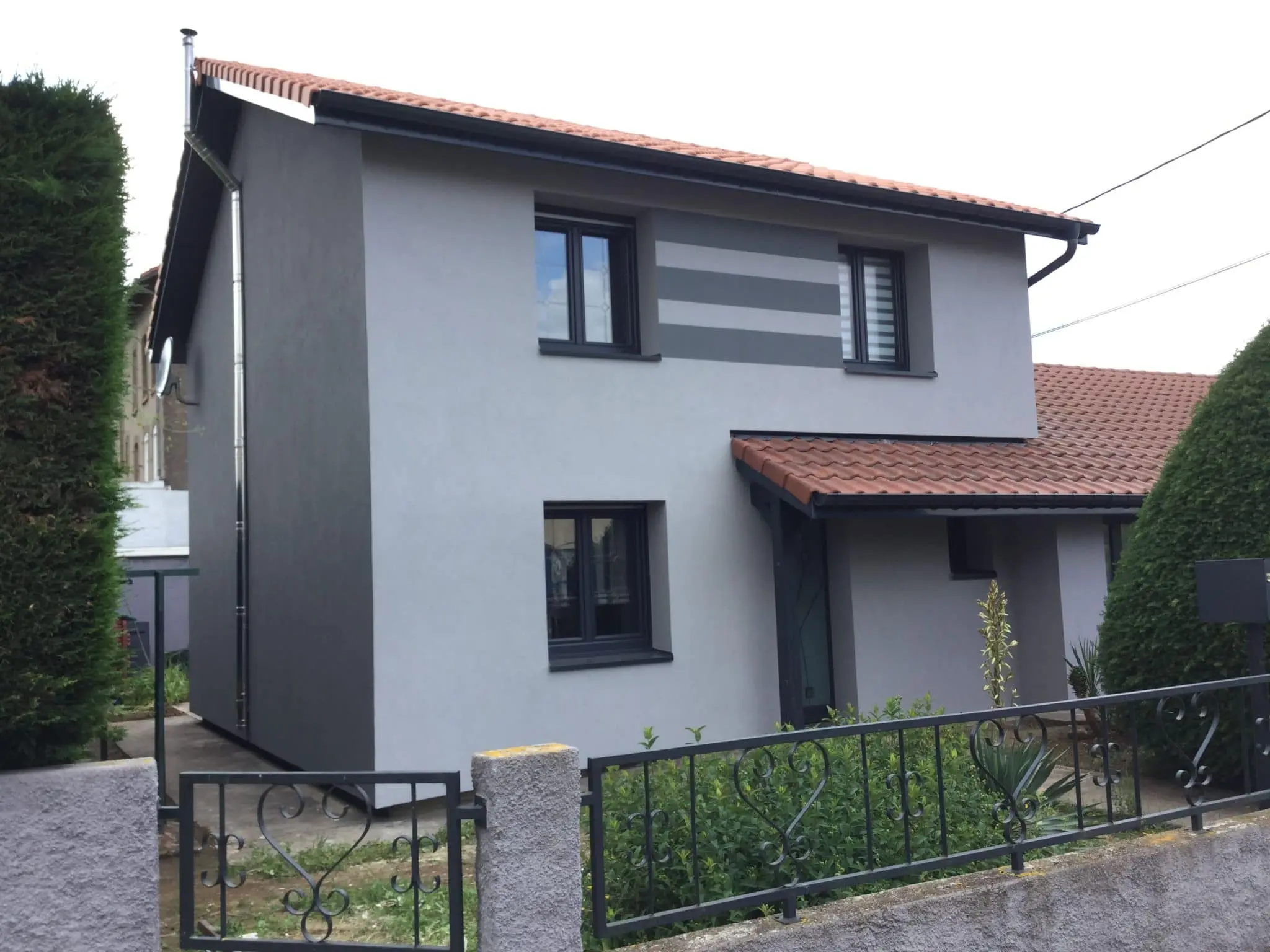 Maison rénovée avec une façade grise moderne, des fenêtres noires et un toit en tuiles rouges, entourée d'un jardin bien entretenu.